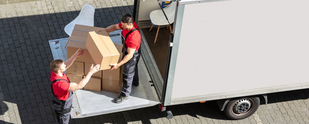 Two movers in red shirts and overalls load boxes onto a moving truck with a lift gate outside, truck door open showing furniture.