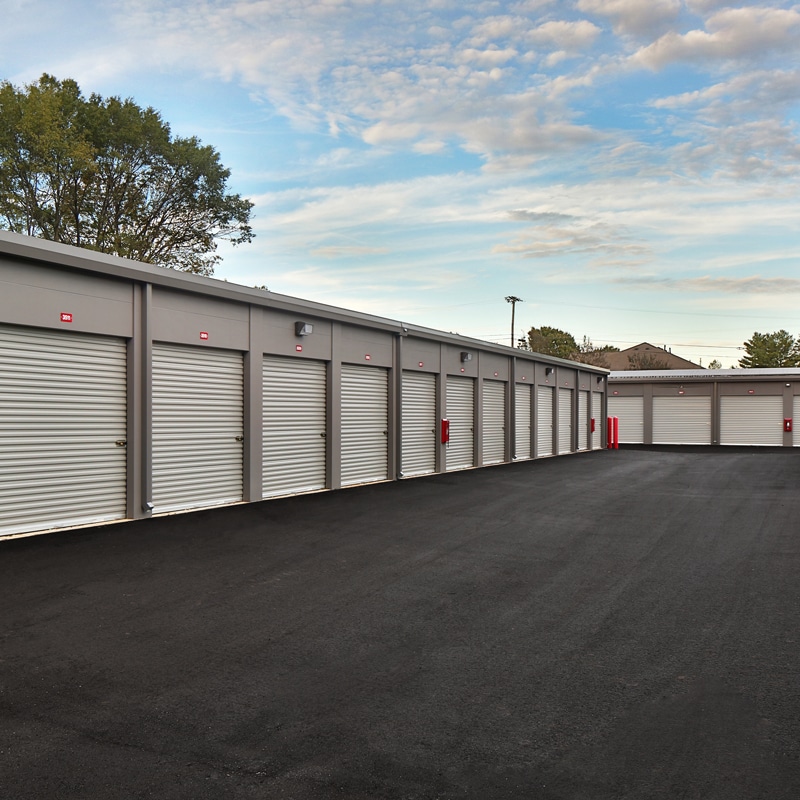 Closed white storage unit doors line a freshly paved lot beneath a partly cloudy sky, trees and rooftops in the background.